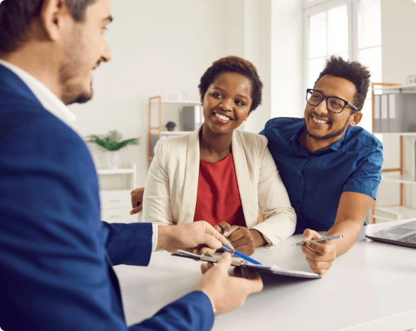 A happy couple sitting in front of an insurance agent who is holding a pen and clipboard full of documents.  