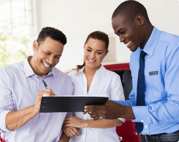 A happy couple signing paperwork on a clipboard held by a happy car salesperson.