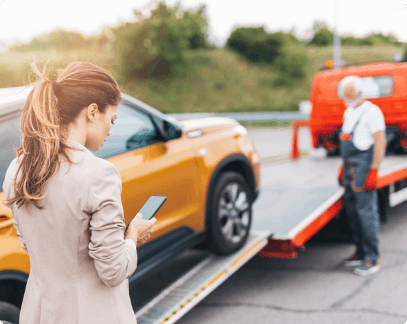 Person standing on the roadside looking at her phone as a tow truck driver loads her car onto his truck.